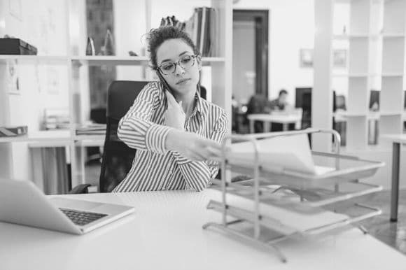 Accounting clerk sitting at her desk, talking on the phone, and putting documents into a desk file.