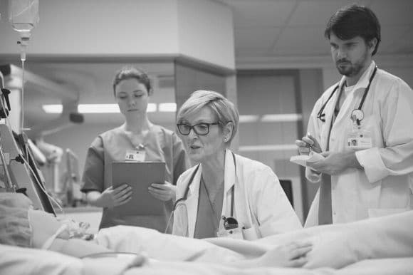 Medical assistant, holding patient clipboard, standing by while physicians consult with a patient.