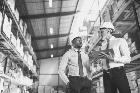 Two warehouse managers, in hard hats and wearing ties, discussing strategy.