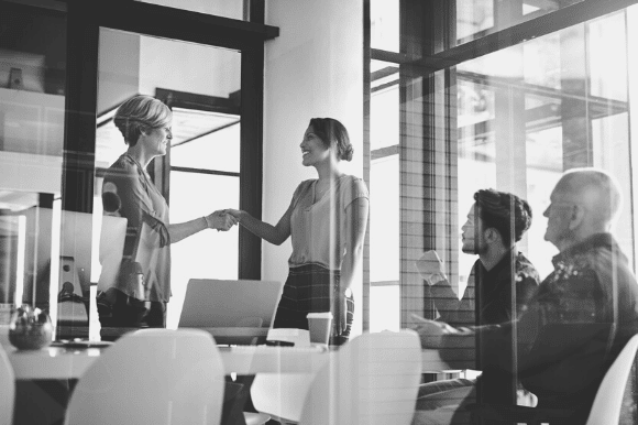 A woman shakes the hand of a job applicant, while colleagues sit nearby at a table, just one part of the recruitment funnel.