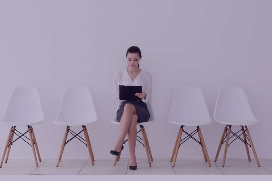 Woman job seeker sits on a chair with an ipad with two empty chairs on either side of her.