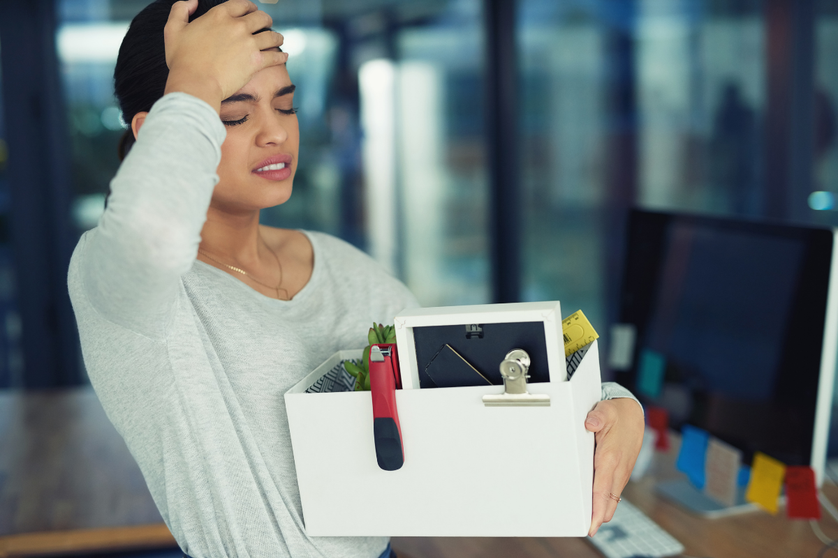 frustrated woman holds a box of office supplies and walks away with her hand on her head.