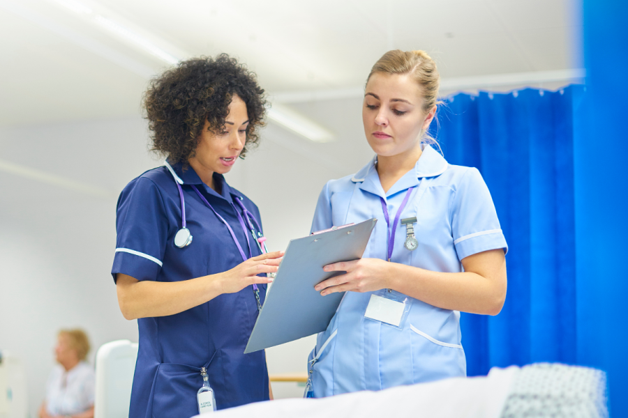 Two female nurses wearing scrubs. One holds a clip board as they discuss patient care.