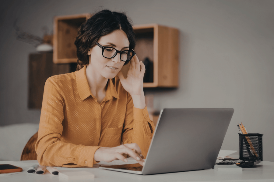 Woman wearing glasses and a yellow blouse sits in front of a laptop and searches for a job.