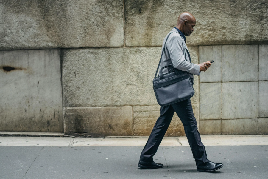 Man in a grey suit and black pants carrying black bag walks on a sidewalk while looking at a cell phone.