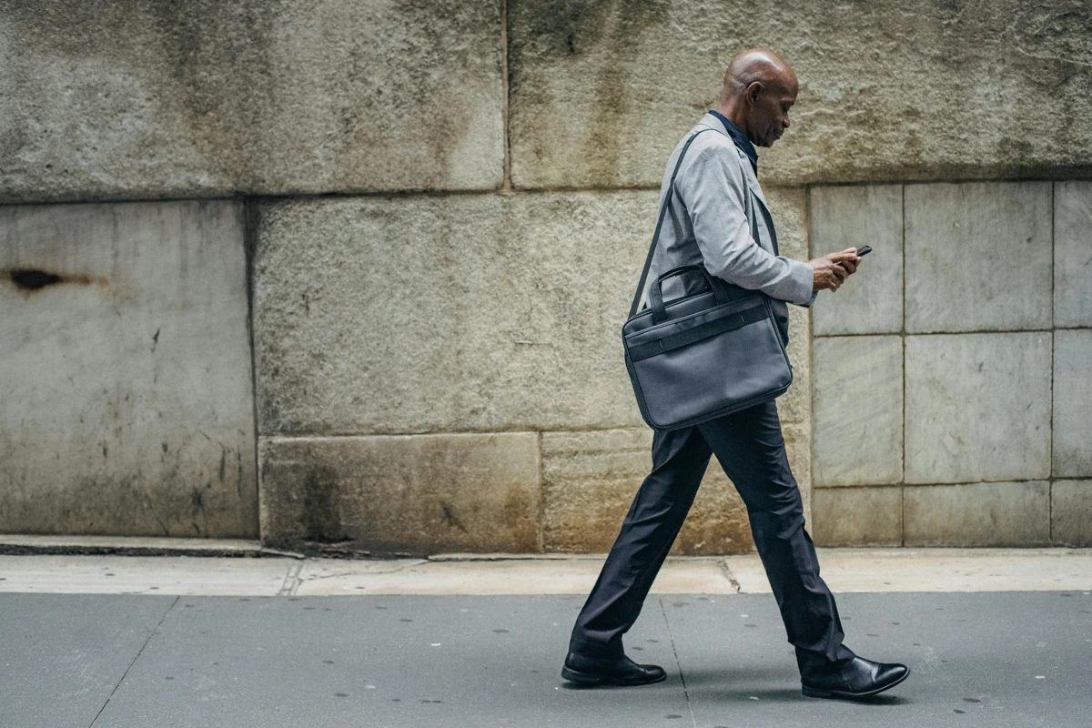 Man in a grey suit and black pants carrying black bag walks on a sidewalk while looking at a cell phone.
