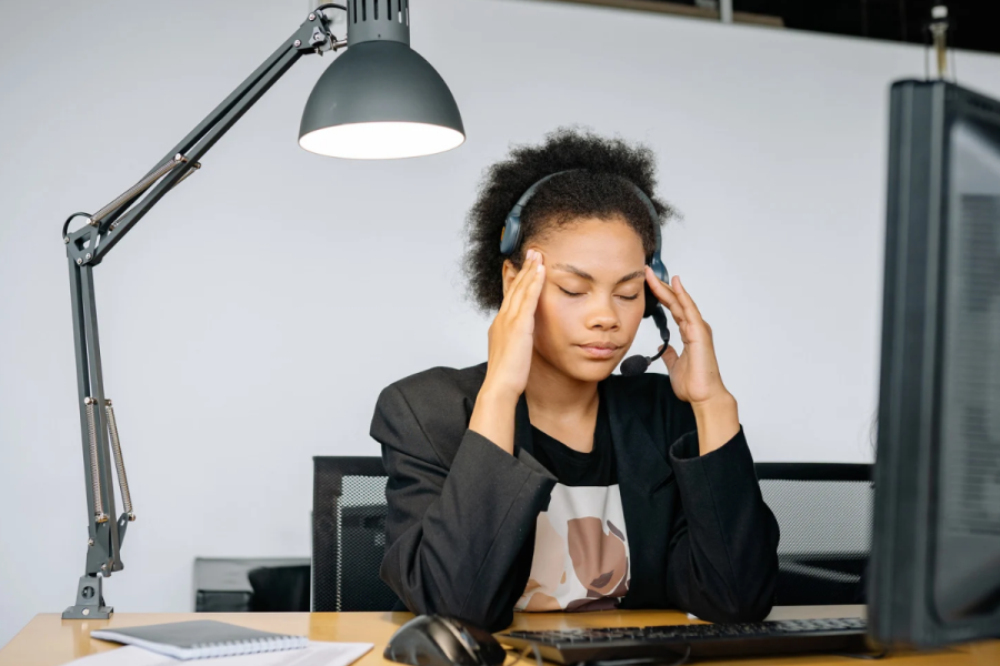 Woman sitting at a desk with a headset on and her eyes closed and her hands on her temples.