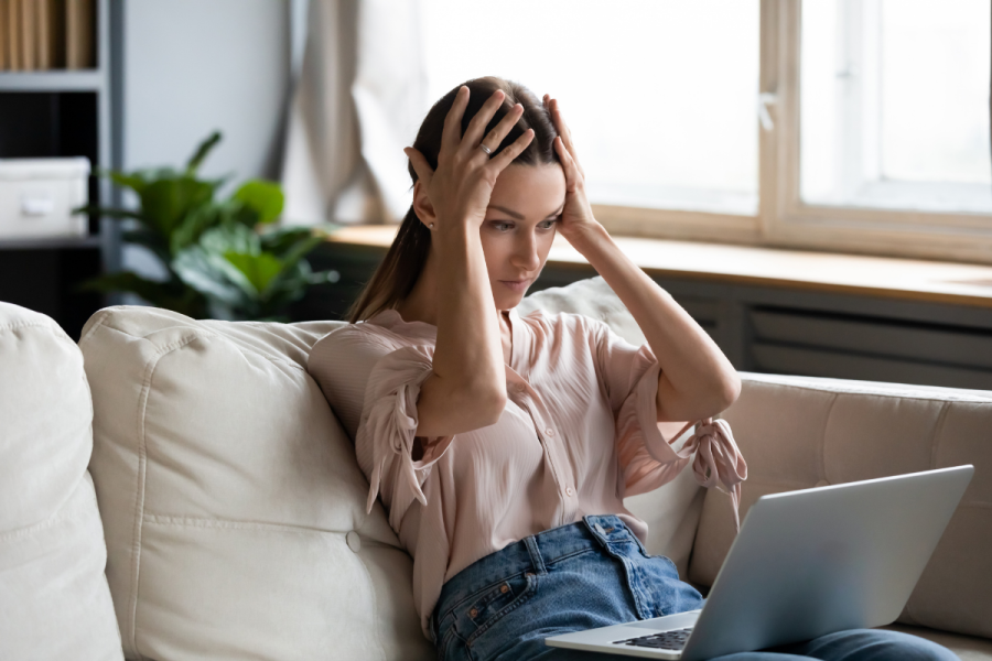 woman with hands on her head in frustration sits in front of her laptop while sitting on the couch