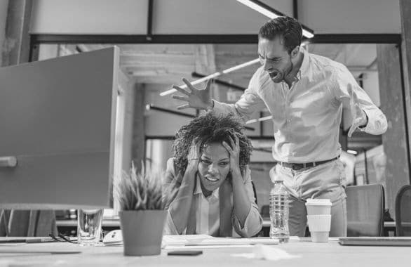 A difficult coworker is harassing his colleague, while she sits with her head in her hands.