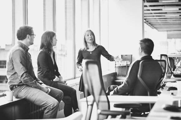 A boss, standing, addressing her seated employees.