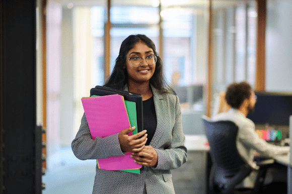 young professional woman in a business suit holding brightly coloured folders, smiling confidently in a modern office environment. She appears to be part of a graduate scheme, with a co-worker visible in the background, working at a desk.