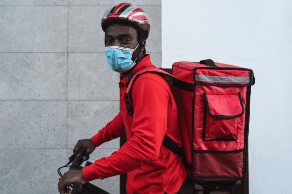 A delivery worker on a bike with a mask looks out at the camera.