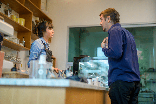 In the image, a man and a woman are engaged in a conversation at a coffee shop counter. The man, dressed in a dark blue jacket, is making a point or asking a question, gesturing with his hand to his chest. The woman, wearing a denim apron typical for a barista, listens attentively with a slight smile, suggesting a friendly or service-oriented interaction. The setting includes typical coffee shop elements like a coffee machine and ceramic wares on shelves, enhancing the casual, everyday life ambiance of the scene, but the man does seem aggressive and unhappy.